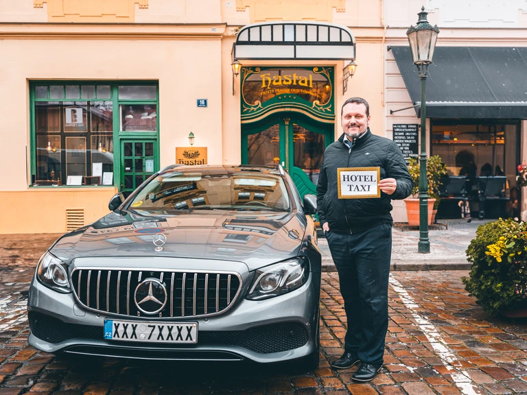 A man in a dark jacket stands next to a silver Mercedes-Benz sedan parked on a cobblestone street in front of the Hotel Haštal. He is holding a small "HOTEL TAXI" sign.